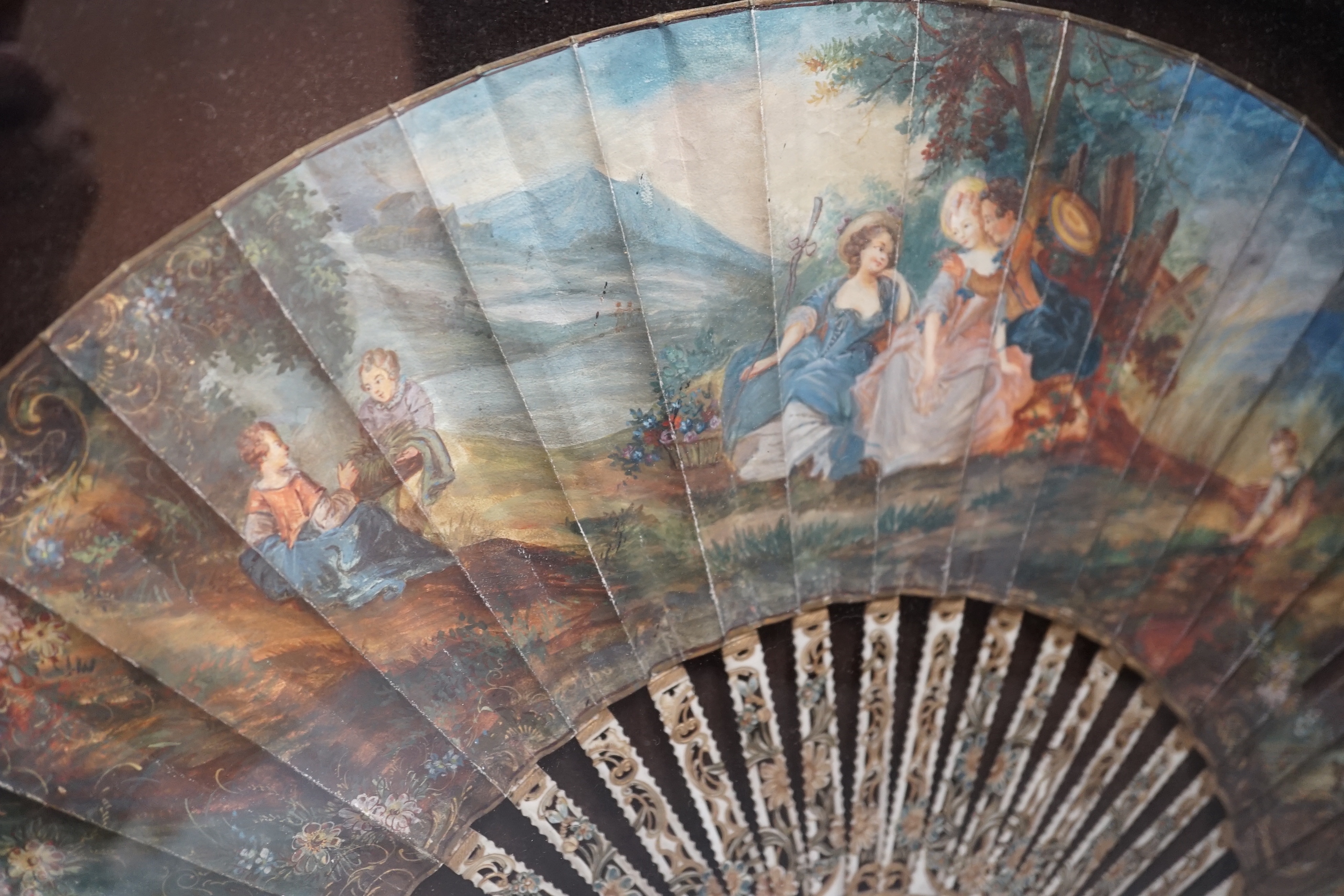 A 19th century French bone fan with pierced decoration, hand painted with figures wearing 18th century dress before a landscape, framed, the frame 62cm wide